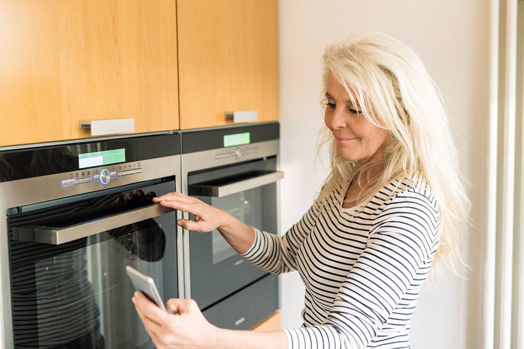 Content mature woman with smartphone checking oven in kitchen of her smart home