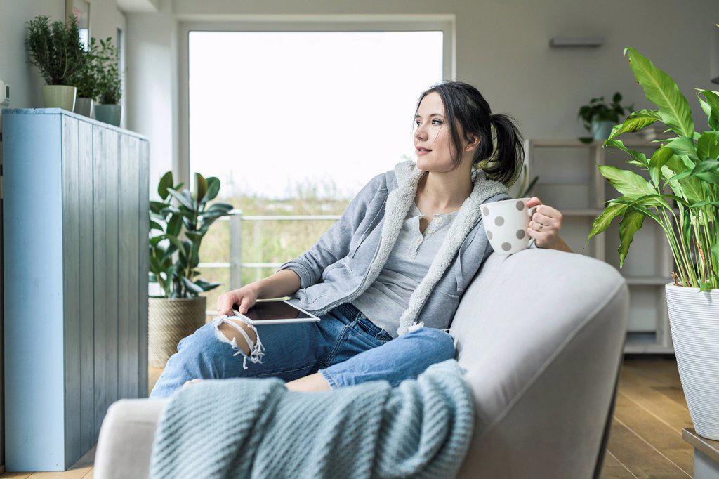 Pensive woman with a mug and tablet sitting on the couch at home