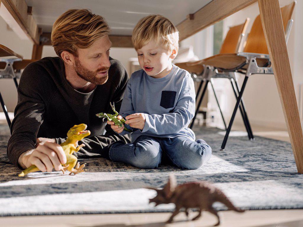Father and son playing with dinosaur figures under the table at home