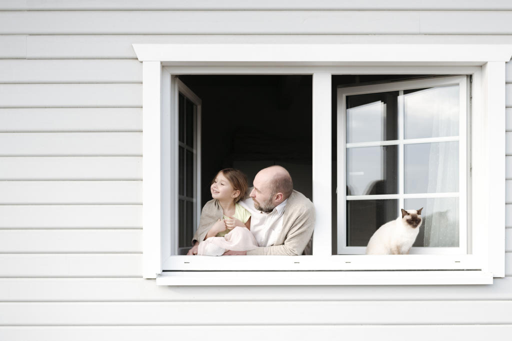 Mature man with smiling daughter and cat leaning out of window of his house