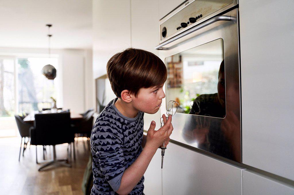 Boy tasting batter while watching his mirror image on surface of oven