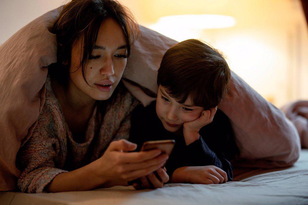 Mother and little son lying together on bed covered with blanket watching movie on smartphone