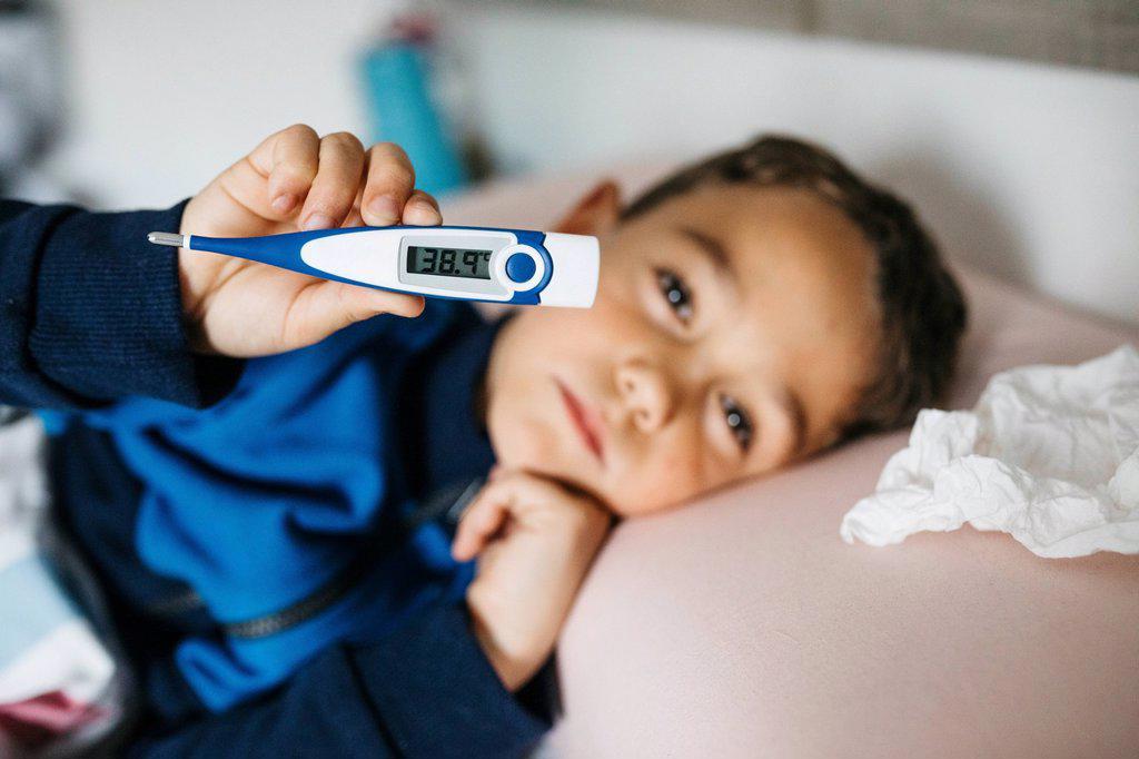 Portrait of sick little boy lying in bed showing digital thermometer
