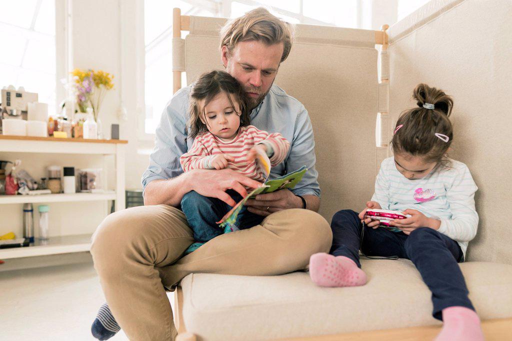 Father reading book to daughter with her sister playing