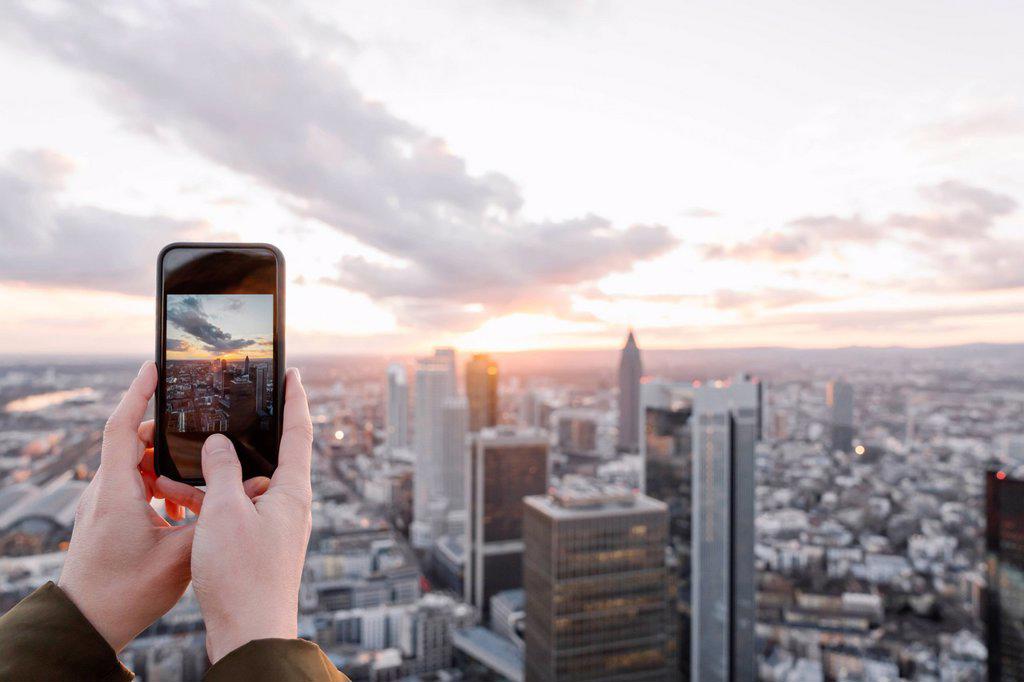 Germany, Hesse, Frankfurt, Hands of woman taking smart phone photos of city downtown at sunset
