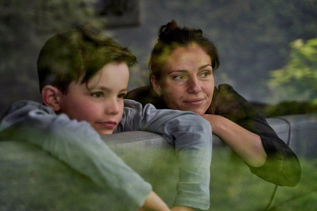 Thoughtful mother and son looking away while relaxing on sofa at home seen through window