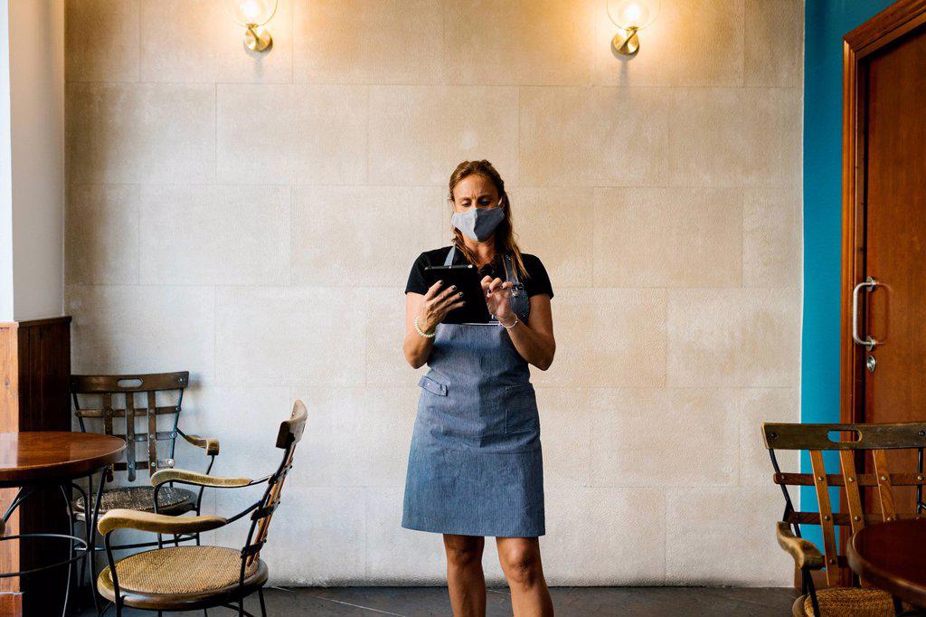 Female cafe owner wearing protective face mask while using digital tablet against wall