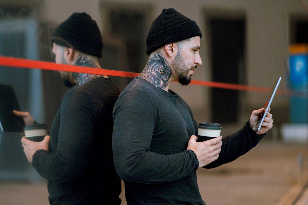 Tattooed hipster man with coffee cup using digital tablet while leaning on glass