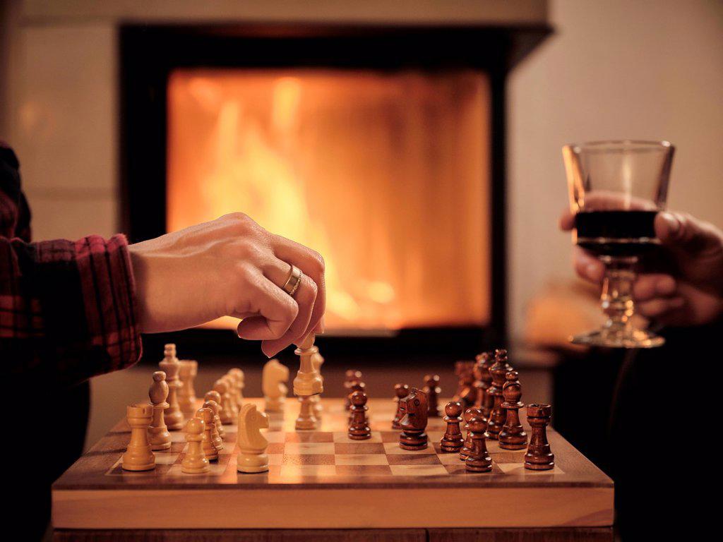 Mature man with wine glass while woman playing chess by fireplace in living room