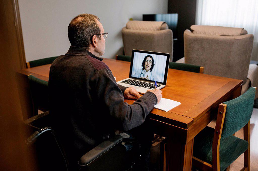 Senior man writing prescription sitting on wheelchair consulted by female doctor through video call at home