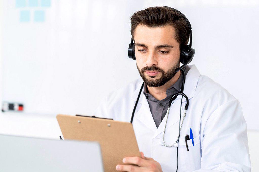 Handsome male doctor wearing headphone while reading report on clipboard in hospital