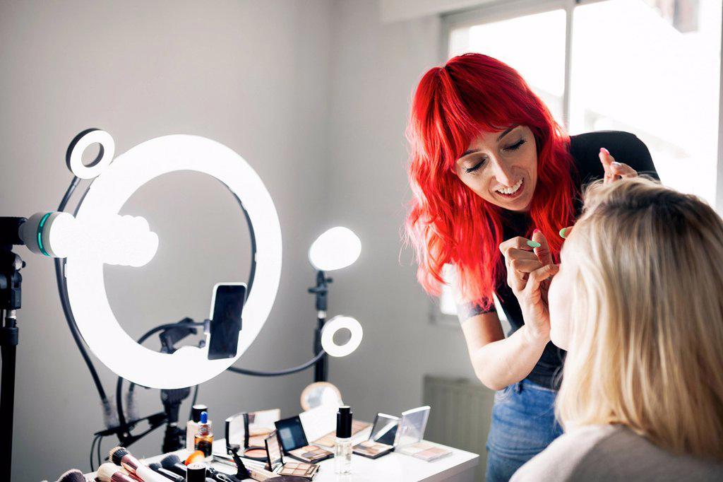 Smiling female make-up artist vlogging through smart phone while applying make-up to model at studio
