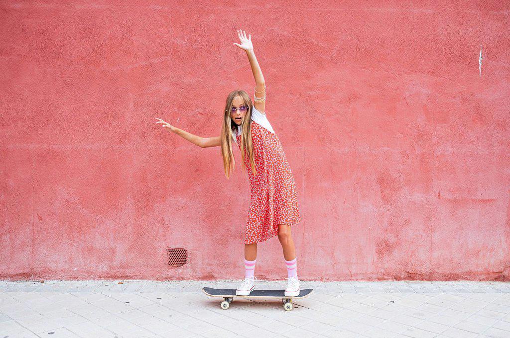 Pre-adolescent girl with hand raised skateboarding on footpath