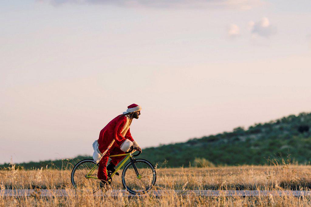 Young man in Santa Claus costume riding bicycle on road during sunset
