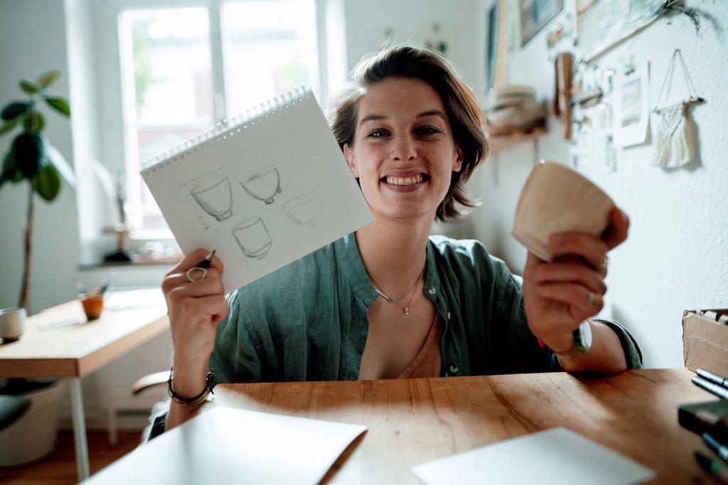 Smiling female illustrator holding spiral notebook and cup on table at home office