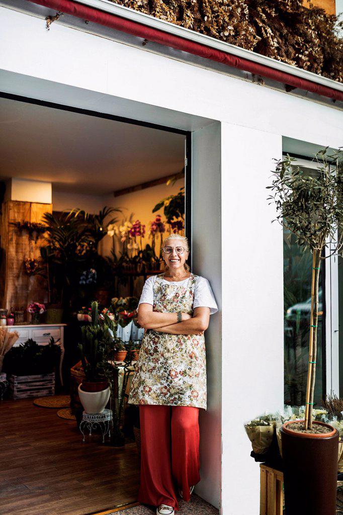 Smiling female florist standing with arms at flower shop entrance
