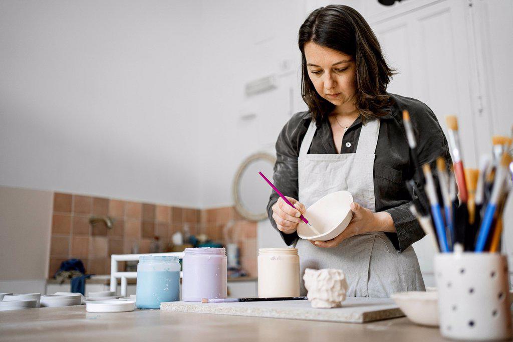 Craftswoman painting ceramic bowl on workbench