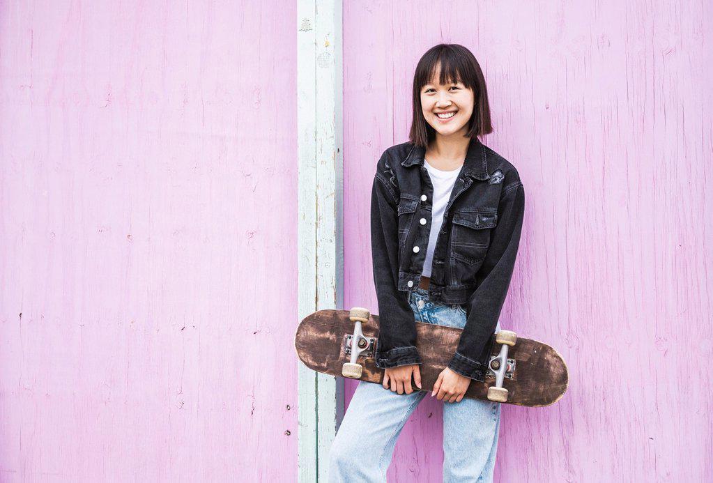Happy teenage girl holding skateboard in front of wall