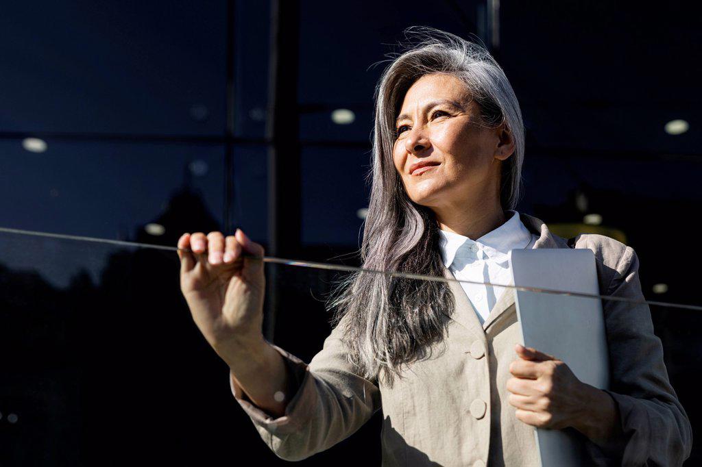 Businesswoman with gray hair holding laptop by glass railing