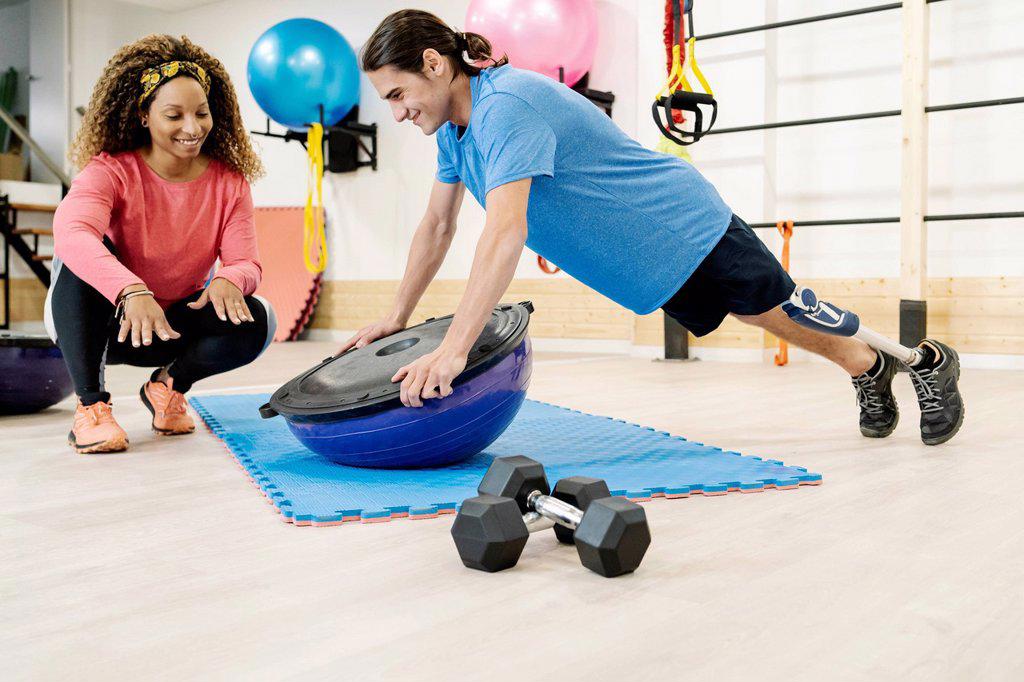 Smiling female instructor assisting disabled man doing push-ups on balance ball in gym