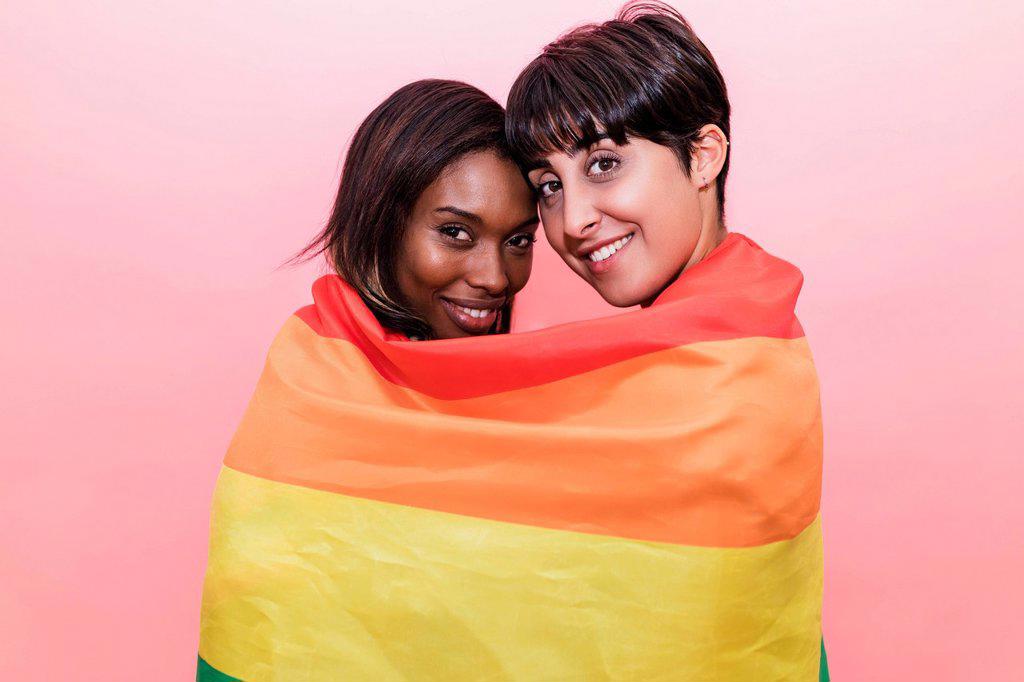 Smiling lesbian couple covered in rainbow flag by pink background