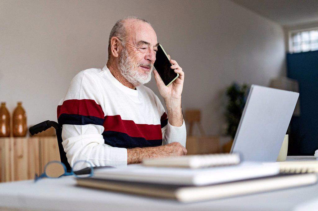 Senior disabled businessman with laptop talking on mobile phone at home