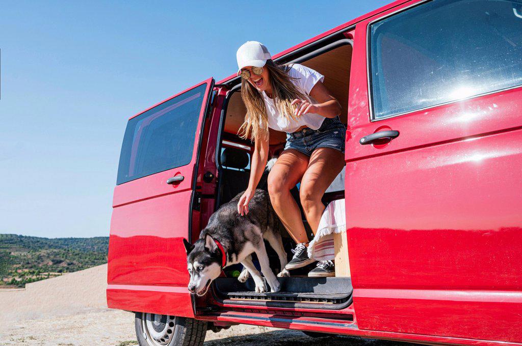 Cheerful woman jumping with dog from red van on sunny day