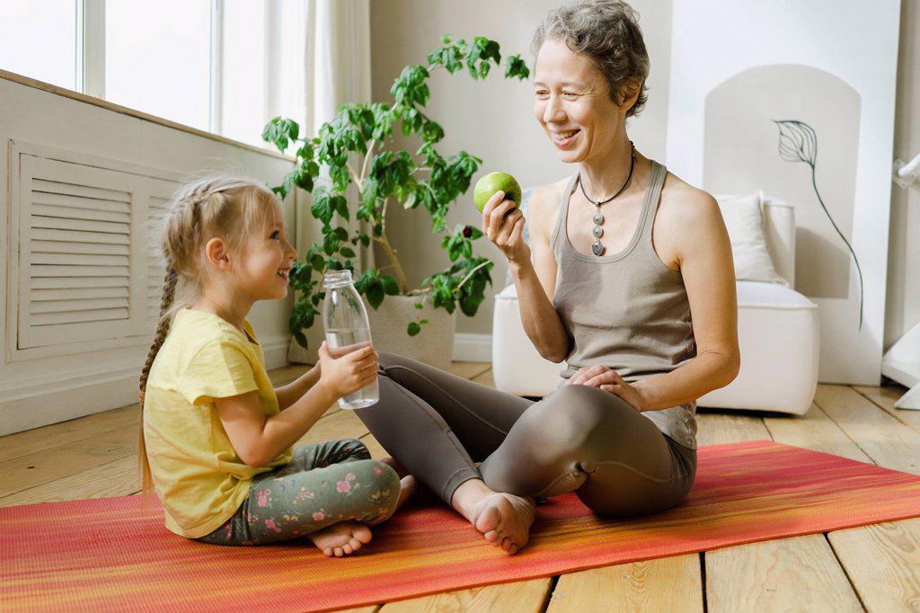 Smiling mother and daughter having food and drink on exercise mat