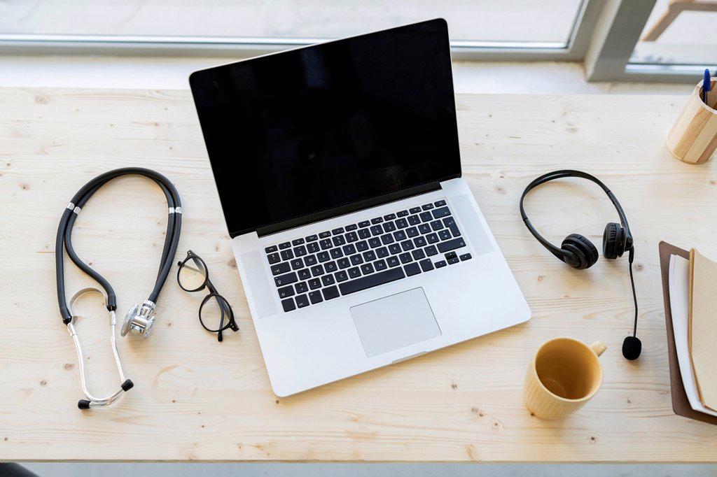 Laptop amidst stethoscope and mug with headphones on desk in home office