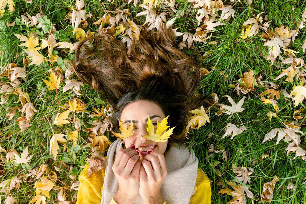 Happy young woman lying on grass holding autumn leaves in park