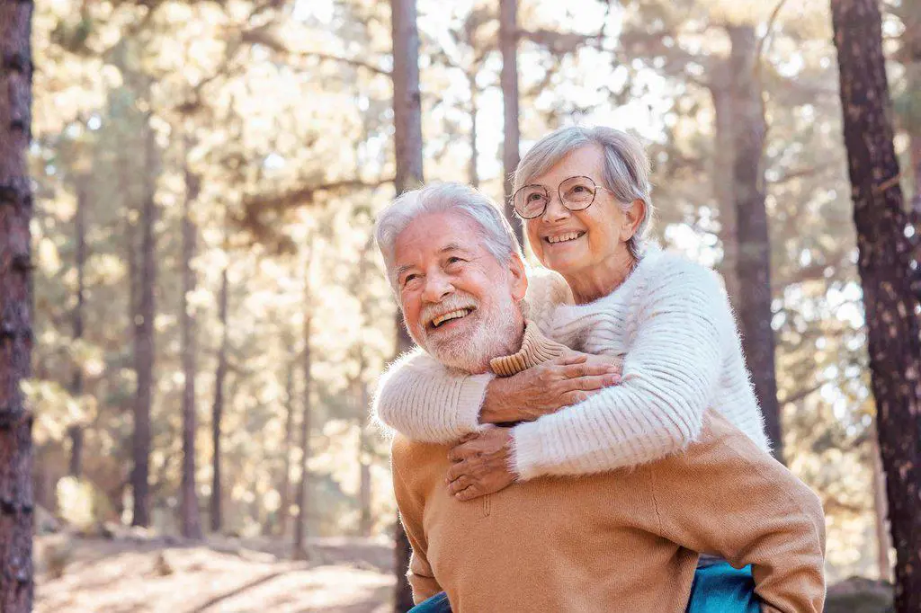 Happy senior man giving piggyback ride to woman wearing eyeglasses in forest