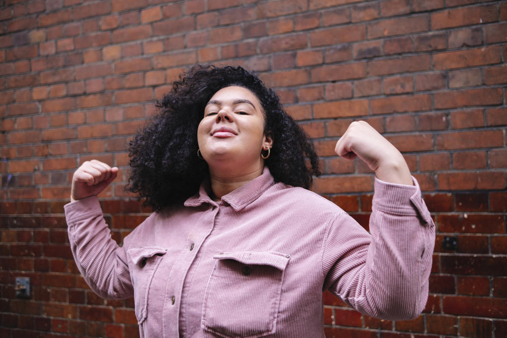 Confident young woman with curly hair flexing muscles in front of brick wall