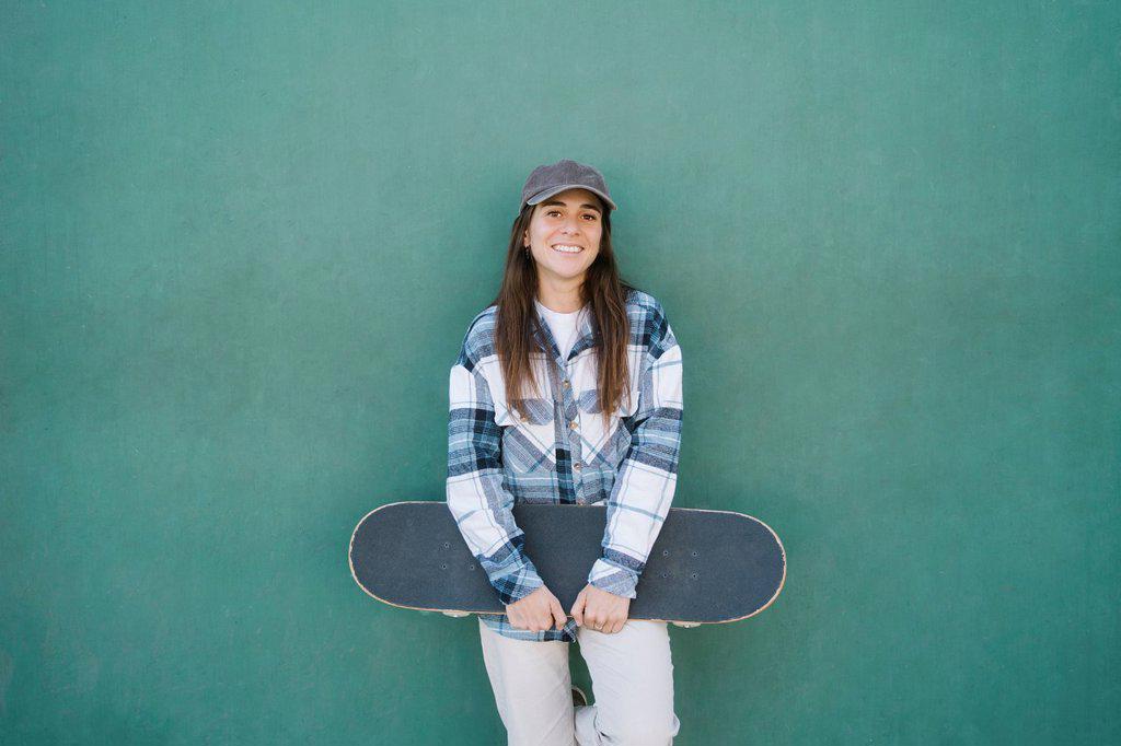 Smiling woman with skateboard standing in front of green wall