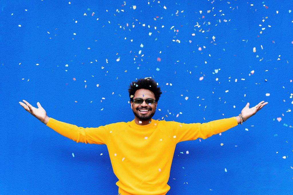 Smiling man with arms outstretched standing under falling confetti in front of blue wall