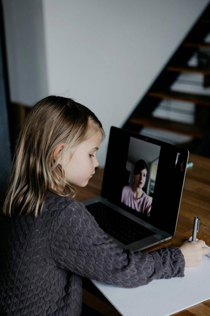 Girl writing on paper studying through video call on laptop at home
