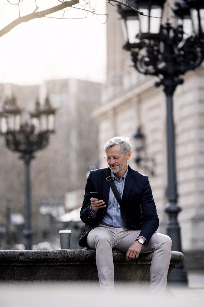 Grey-haired businessman in the city with smartphone and coffee to go