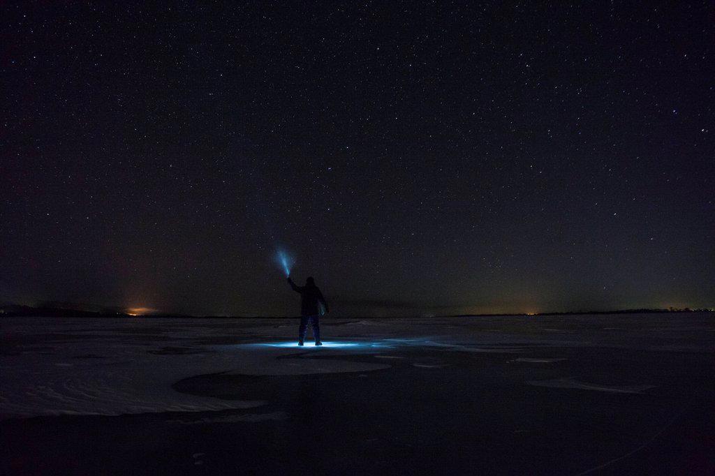 Russia, Amur Oblast, silhouette of man with blue ray standing on frozen Zeya River at night under starry sky