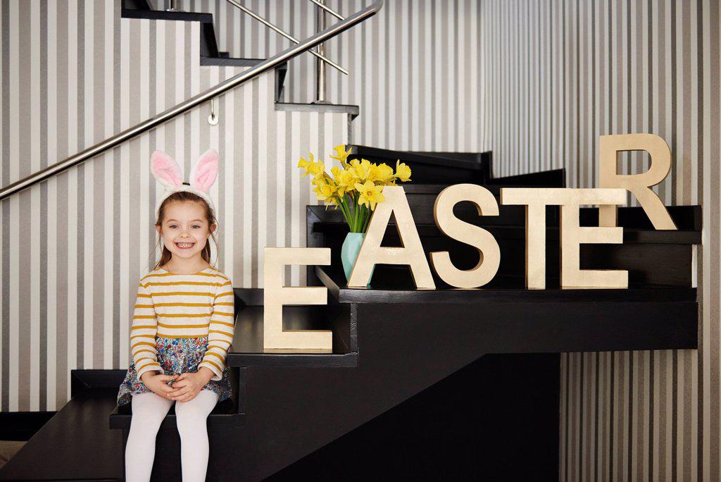 Portrait of smiling girl with bunny ears sitting on stairs next to word 'Easter'