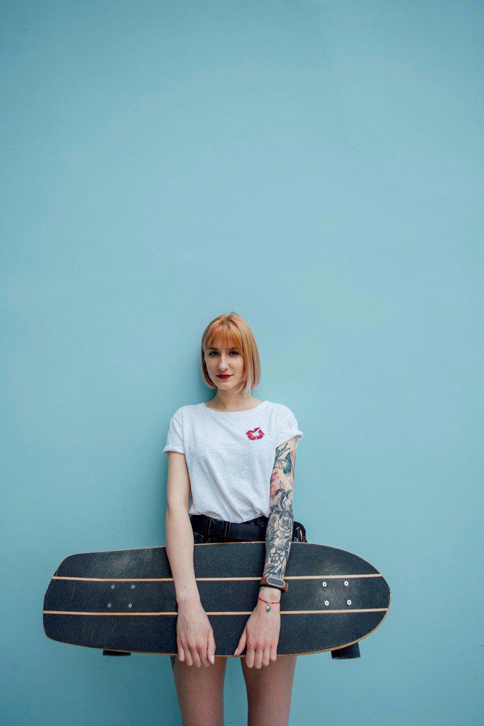 Portrait of cool young woman holding carver skateboard standing at turquoise wall
