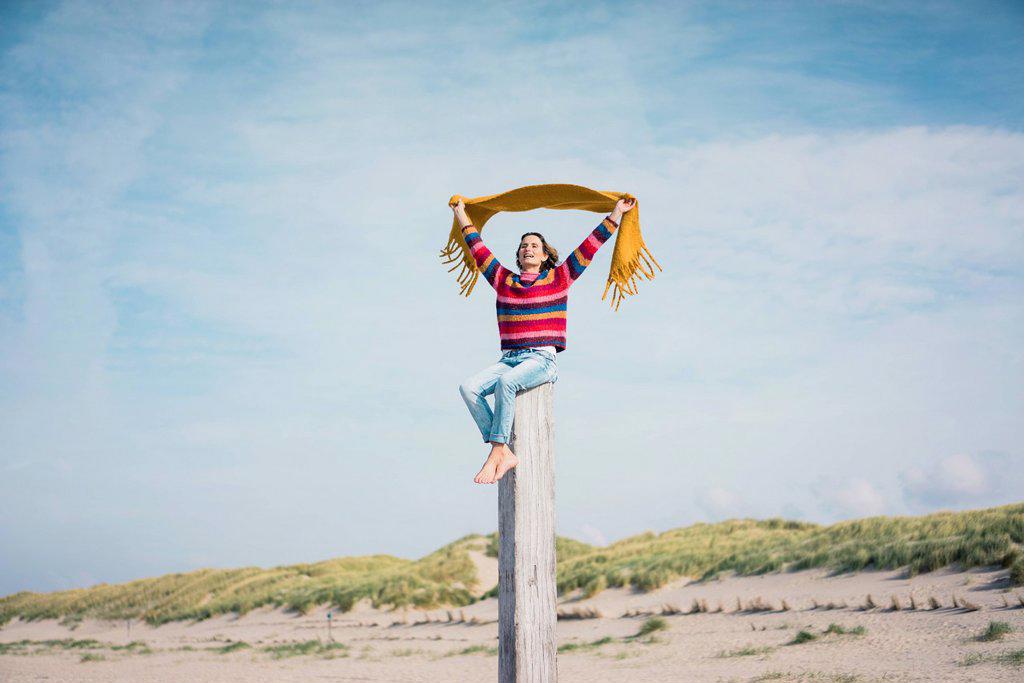 Mature woman sitting on a wood pole on the beach, holding scarf in the wind