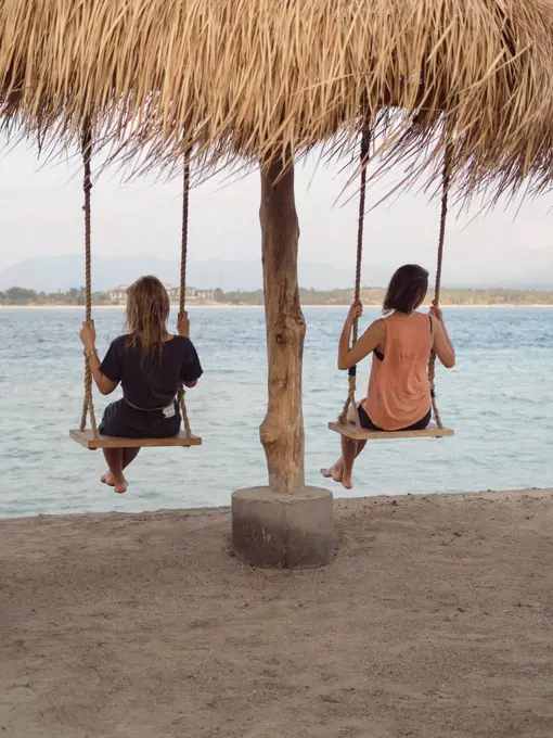 Back view of two women sitting on swings at seafront, Gili Islands, Bali