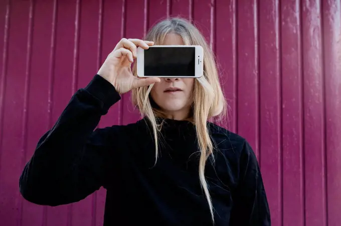 Portrait of blond young woman covering eyes with smartphone