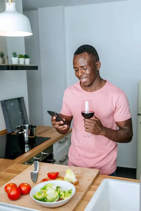 Smiling young man with wine glass using cell phone while preparing salad