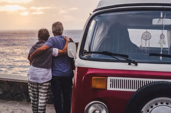 Senior couple traveling in a vintage van, looking at the sea with arms around
