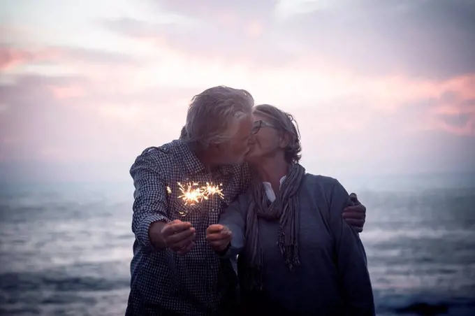 Kissing senior couple standing in front of the sea by sunset holding sparklers