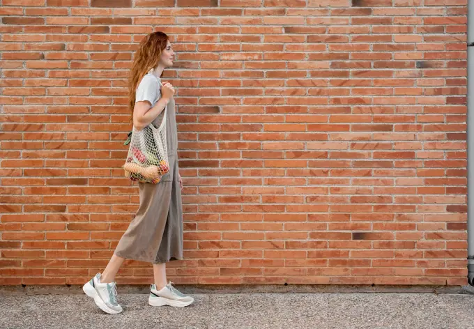 Young woman carrying groceries in mesh bag while walking on footpath by brick wall