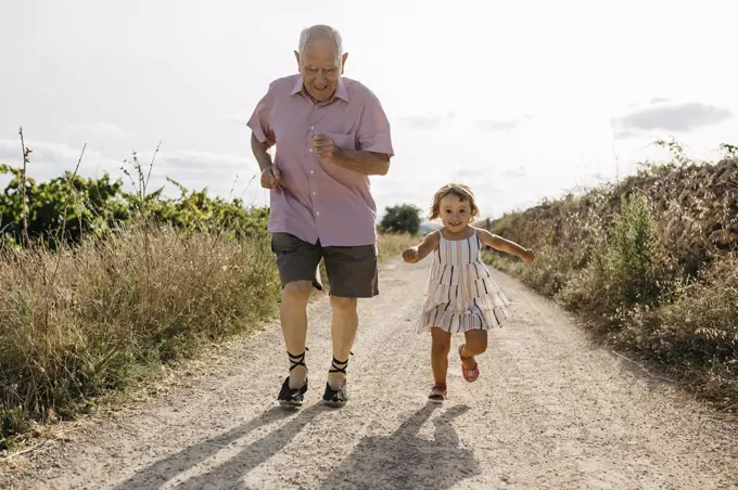 Playful senior man running with granddaughter on dirt road amidst plants