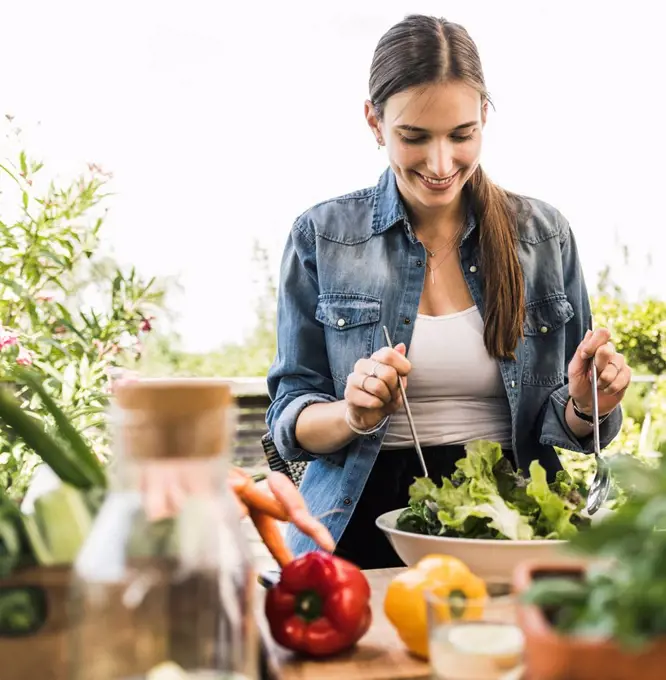 Smiling young woman making salad in bowl while sitting against clear sky at yard