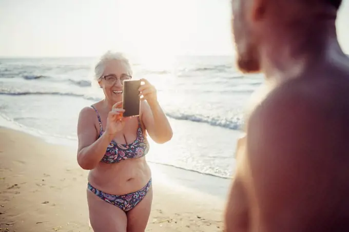 Senior woman in bikini taking photo of man while standing at beach