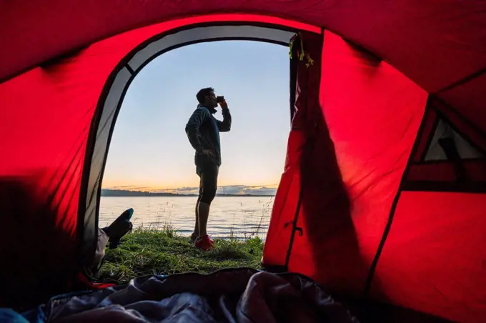 Man camping in Estonia, standing in front of tent, watching sunset